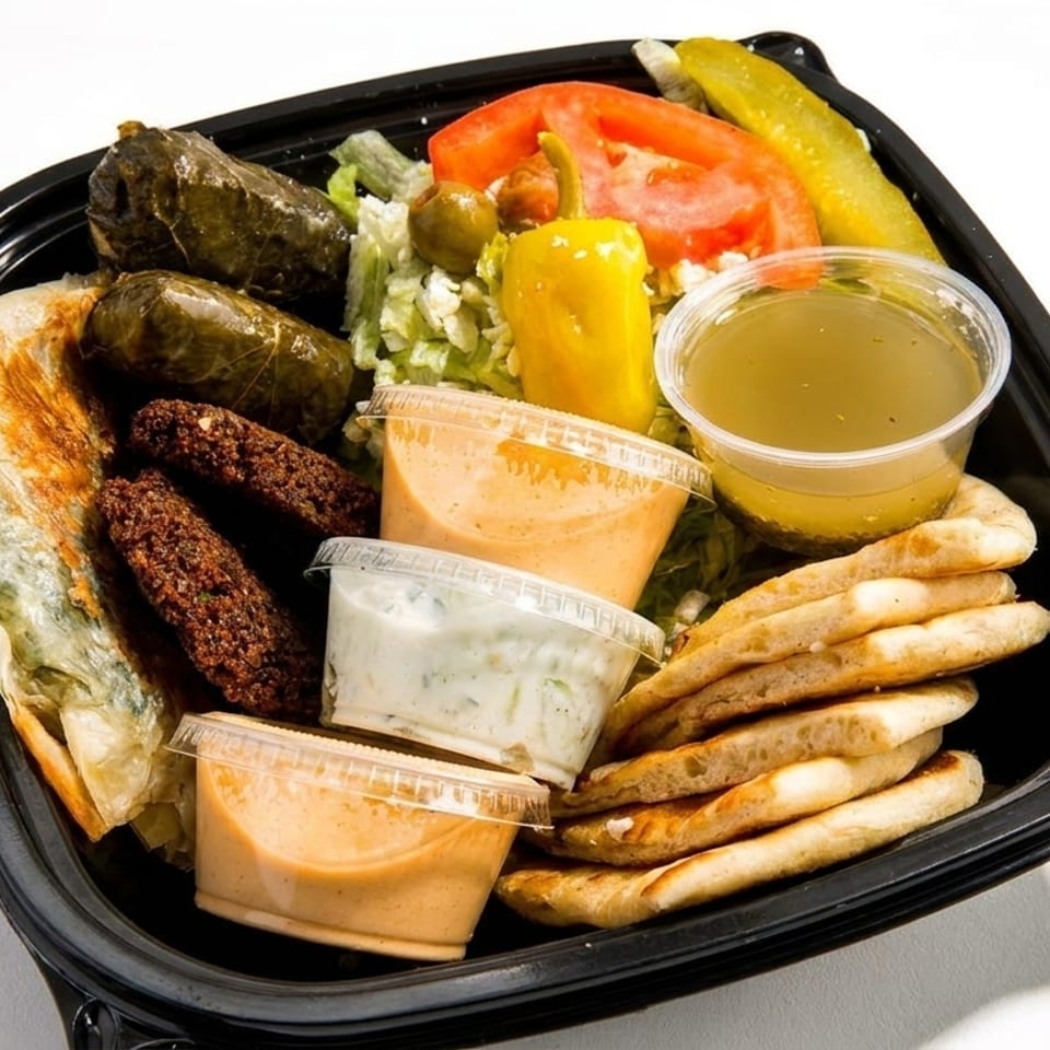 A black takeout container filled with falafel, stuffed grape leaves, salad, pita bread, and sauces.