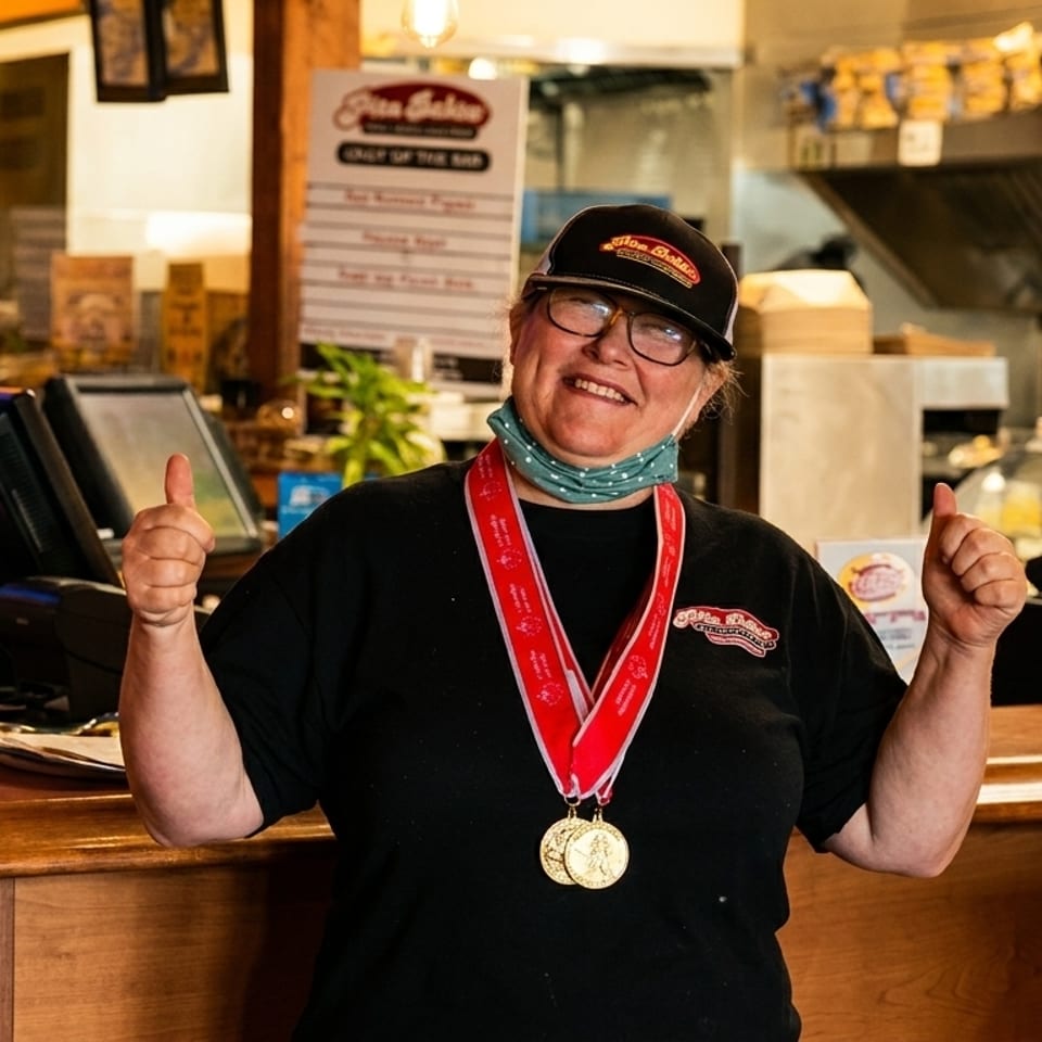 Smiling woman in a black shirt and hat, wearing medals, giving two thumbs up behind a counter.