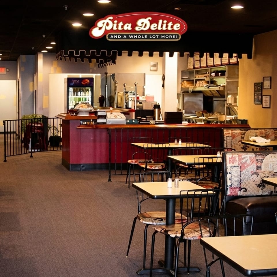 Interior of Pita Delite restaurant with tables, chairs, booths, and a service counter under the brand sign.